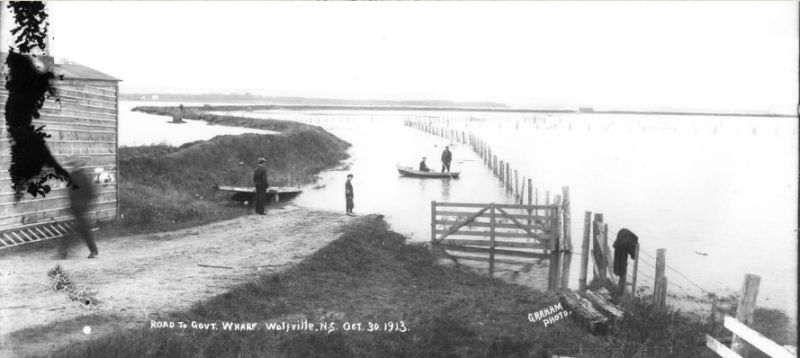 Black and white photo with handwritten words on it: "ROAD TO GOVT. WHARF. Wolfville, N.S. Oct 30 1913." showing a scene of flooded dyke lands.