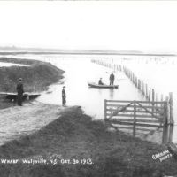 Black and white photo with handwritten words on it: "ROAD TO GOVT. WHARF. Wolfville, N.S. Oct 30 1913." showing a scene of flooded dyke lands.